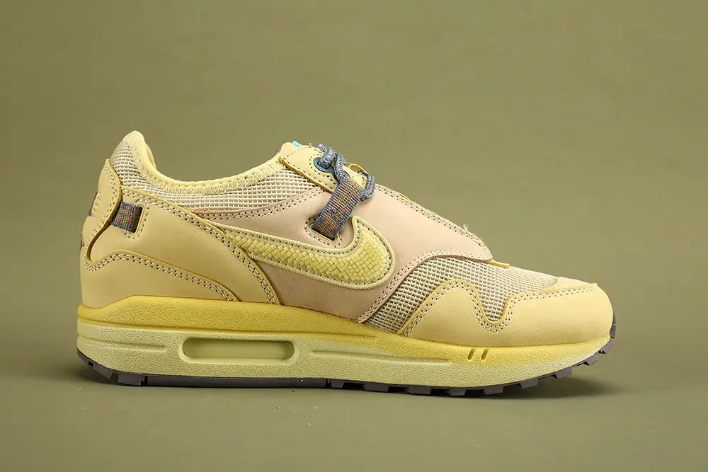 Air Max 1 Cactus Jack Saturn Gold Tea Tree Mist - Toe Box Close Up on Olive Background Showing Cream Coarse Square-Grid Woven Mesh Toe Box Body Saturn Gold Smooth Nubuck Toe Box Overlay with Yellow Tonal Edge Stitching Multicolour Reflective Woven Webbing Lace Loops Grey Rope Laces with Multicolour Speckle and Saturn Gold AM1 Midsole with Saturn Gold Suede Mudguard and Dark Grey Rubber Outsole
