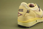 Air Max 1 Cactus Jack Saturn Gold Tea Tree Mist - Rear View of Both Shoes on Olive Background Showing Mauve Purple-Brown Triangular Pennant Flag Patches at Heel Counter with Co-Branded Text and Swoosh on Left and CACTUS JACK Text on Right Multicolour Reflective Woven Webbing Heel Pull Tabs Saturn Gold Nubuck Heel Counter Overlay and Saturn Gold AM1 Midsole