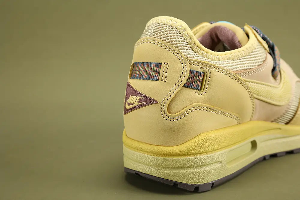 Air Max 1 Cactus Jack Saturn Gold Tea Tree Mist - Rear View of Both Shoes on Olive Background Showing Mauve Purple-Brown Triangular Pennant Flag Patches at Heel Counter with Co-Branded Text and Swoosh on Left and CACTUS JACK Text on Right Multicolour Reflective Woven Webbing Heel Pull Tabs Saturn Gold Nubuck Heel Counter Overlay and Saturn Gold AM1 Midsole