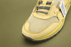 Air Max 1 Cactus Jack Saturn Gold Tea Tree Mist - Both Shoes Three-Quarter View on Olive Background Showing Saturn Gold Woven-Texture Swoosh Cream Coarse Woven Mesh Upper Saturn Gold Nubuck Overlays Multicolour Reflective Woven Webbing Lace Loops and Saturn Gold AM1 Midsole with Heel Air Window