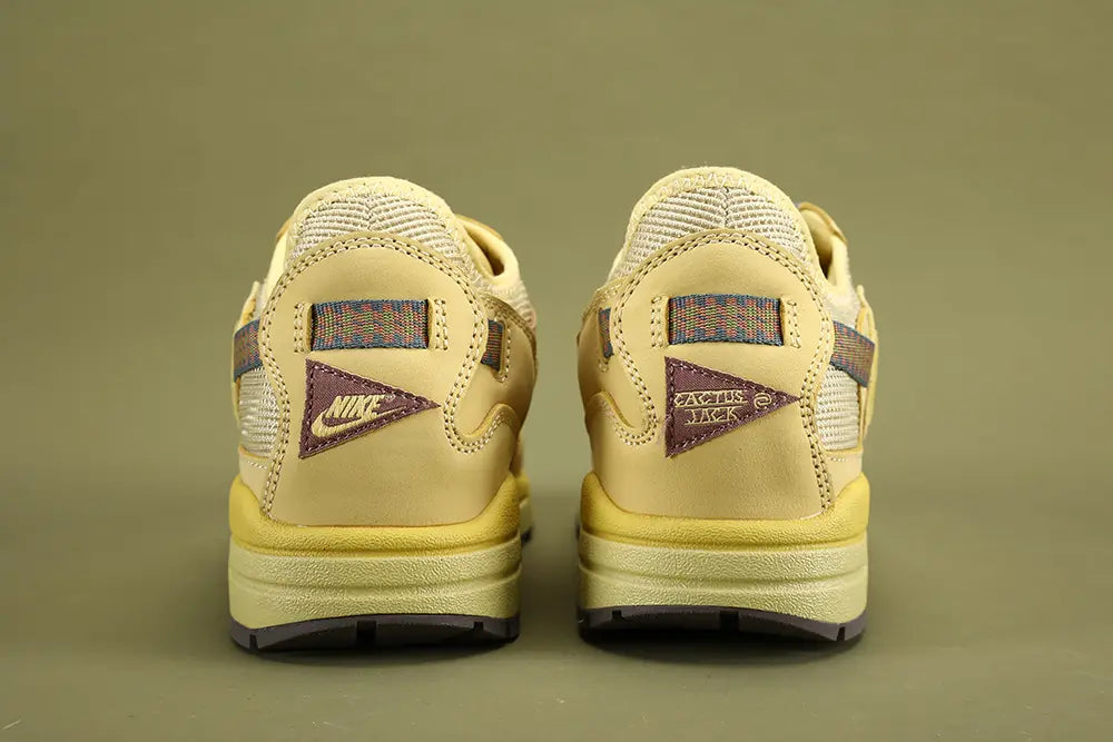 Air Max 1 Cactus Jack Saturn Gold Tea Tree Mist - Full Lateral Single Shoe View on Olive Background Showing Complete Lateral Profile Saturn Gold Woven Swoosh Cream Woven Mesh Multicolour Webbing Lace Loops Teal Lace Lock White Hang Tag Saturn Gold Suede Mudguard and Saturn Gold AM1 Midsole with Dark Grey Outsole