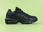 Air Max 95 Essential Triple Black - Outsole View on Green Background Showing Full Black Rubber AM95 Square Block Waffle Tread with Central Translucent Window Revealing Black White Striped Insole