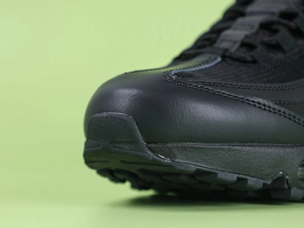 Air Max 95 Essential Triple Black - Heel Lateral Close Up on Green Background Showing Black Snake Embossed Leather Panel with Tonal Black Embroidered Swoosh and Black Mesh Quarter Panel Detail