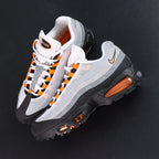 Air Max 95 OG Bright Mandarin 2.0 2025 - Pair Three Quarter Lateral View with Pikachu on Sofa Showing Full Upper Black Suede Toe Box Grey Gradient Spine Orange Mini Swoosh Orange Lace Loops and Orange Air Units