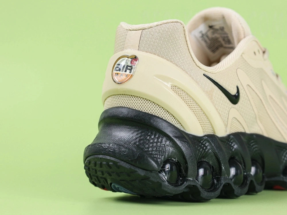 Air Max Dn8 Desert Khaki Black - Tongue Close Up on Green Background Showing White Oval airMAX Rubber Badge with Black Border and Black Lettering on Bone Mesh Tongue