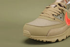 Off-White x Air Max 90 Desert Ore - Tongue and Lace Area Close Up on Olive Background Showing White Woven NIKE SPORTSWEAR Label with Orange Swoosh Metallic Gold Shimmer Tongue Lining and Hot Coral Overlock Stitching on Suede Overlays