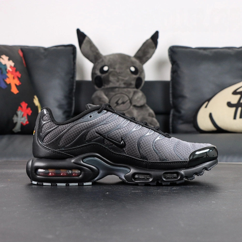 Paris Saint-Germain x Air Max Plus Black Particle Grey - Rear Heel View of Both Shoes on Dark Background Showing Yellow Tn AIR Heel Tab Badge with Red Circular Accent Dot and Full Black AM Plus Outsole