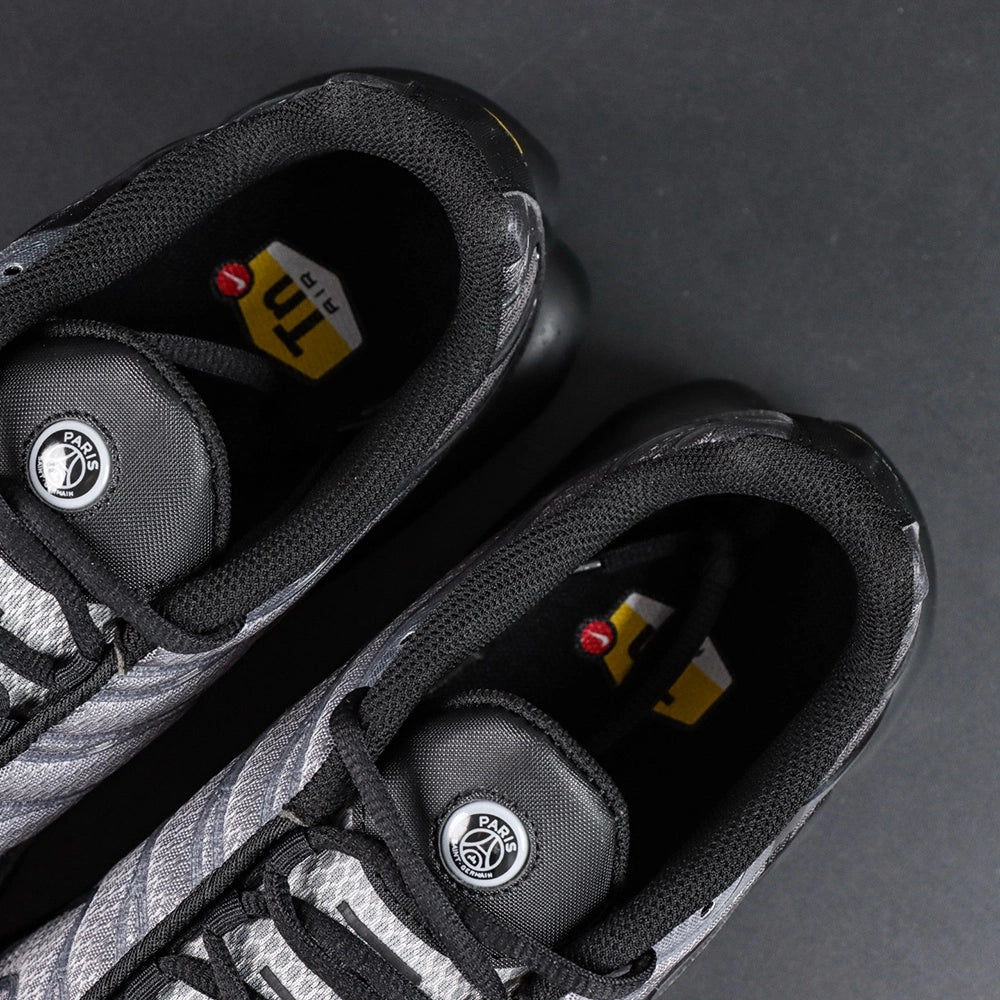 Paris Saint-Germain x Air Max Plus Black Particle Grey - Three-Quarter Lateral View of Both Shoes on Dark Background Showing Full Black and Particle Grey Upper Construction Large PSG Lettering on Hex Mesh and Black TN Midsole