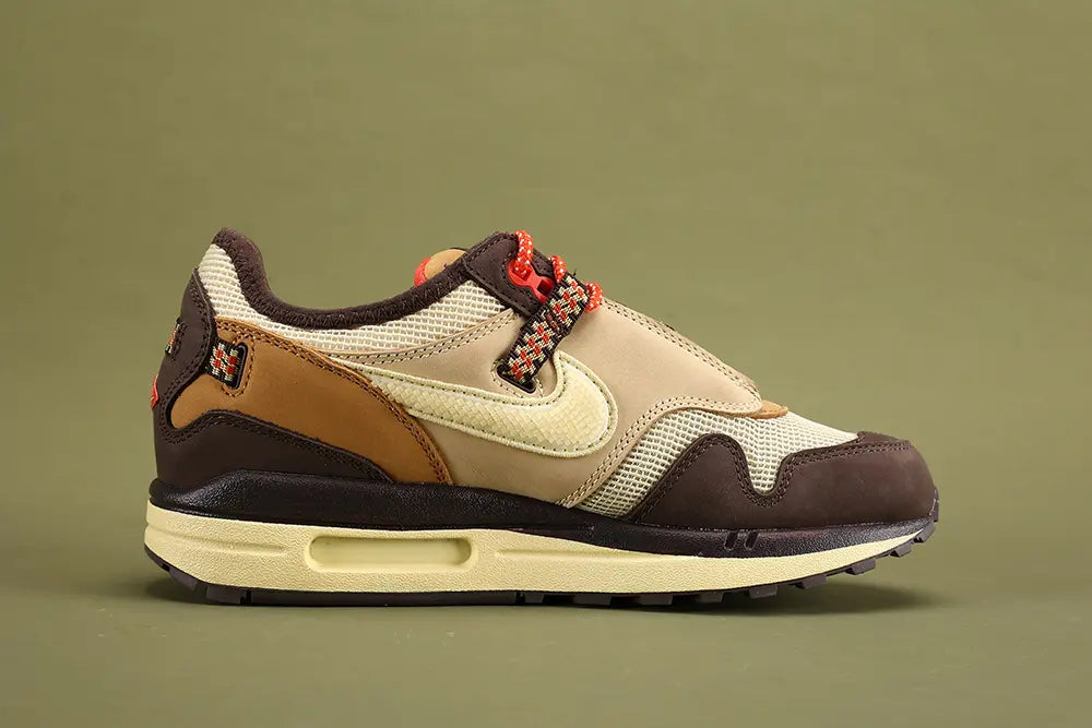 Air Max 1 Cactus Jack Baroque Brown Wheat Cream University Red - Lace Area Close Up on Olive Background Showing Cream Circular Lace Lock with Cream Logo Badge Black Multicolour Stripe Woven Webbing Lace Loops University Red White-Speckle Rope Laces Wheat Quarter Panel and University Red Collar Lining