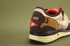 Air Max 1 Cactus Jack Baroque Brown Wheat Cream University Red - Both Shoes Three-Quarter Lateral View on Olive Background Showing Full Upper Profile Cream Woven Swoosh Baroque Brown Nubuck Cream Woven Mesh Wheat Quarter Panel Black Webbing Lace Loops University Red Rope Laces White MADE IN VIETNAM Hang Tag and Cream AM1 Midsole with Heel Air Window