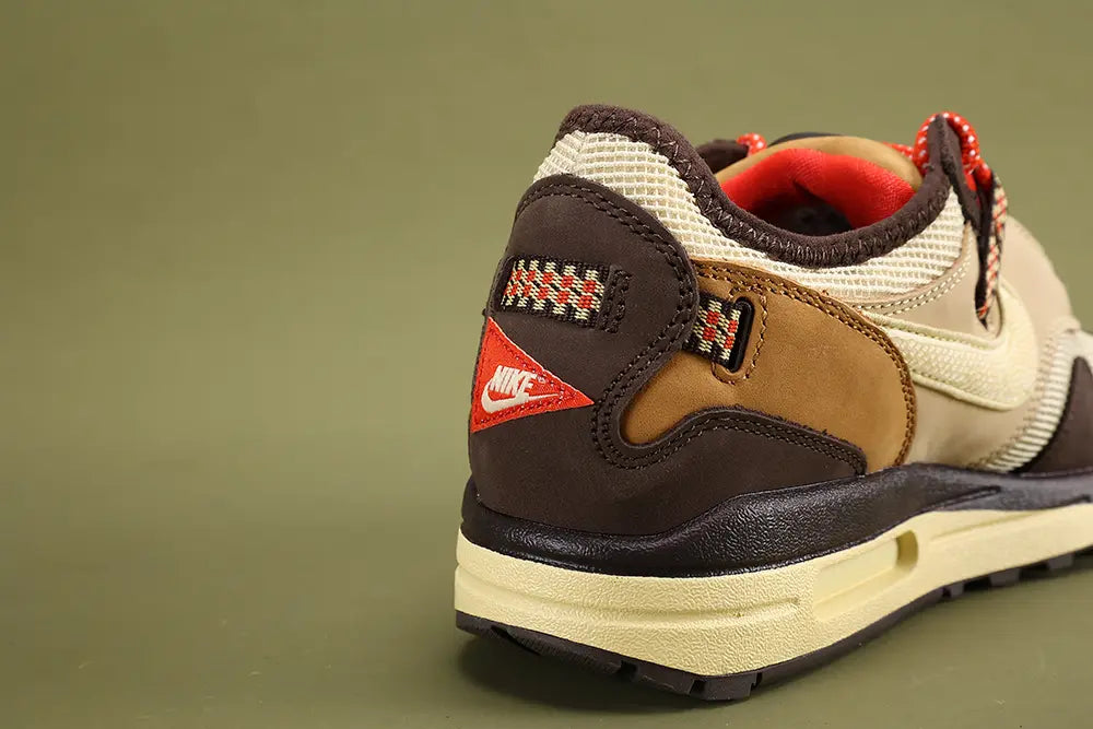 Air Max 1 Cactus Jack Baroque Brown Wheat Cream University Red - Both Shoes Three-Quarter Lateral View on Olive Background Showing Full Upper Profile Cream Woven Swoosh Baroque Brown Nubuck Cream Woven Mesh Wheat Quarter Panel Black Webbing Lace Loops University Red Rope Laces White MADE IN VIETNAM Hang Tag and Cream AM1 Midsole with Heel Air Window