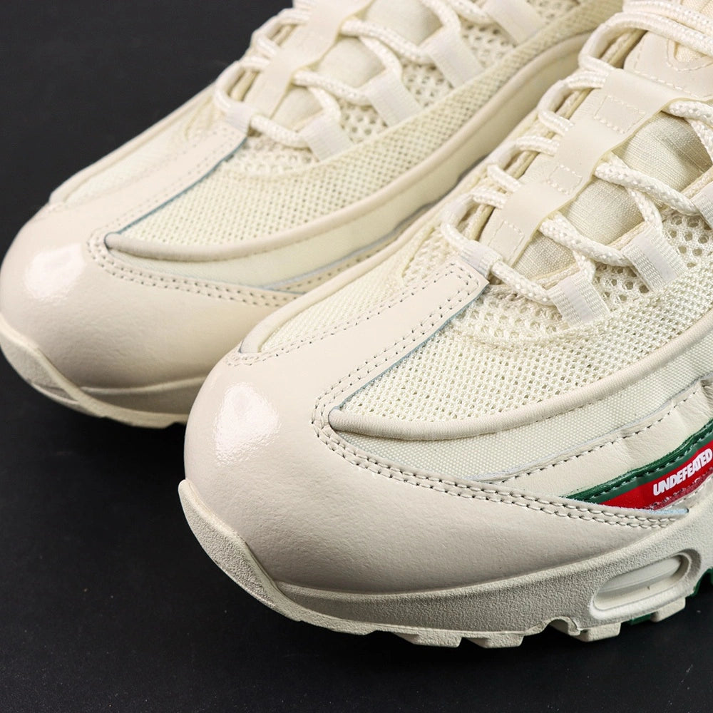 Undefeated x Air Max 95 Sail 2025 - Three-Quarter Lateral Top View of Both Shoes on Dark Background Showing Sail AM95 Upper Dark Green and Red UNDEFEATED Dual Stripe on Lateral Midfoot and Sail White Rope Laces