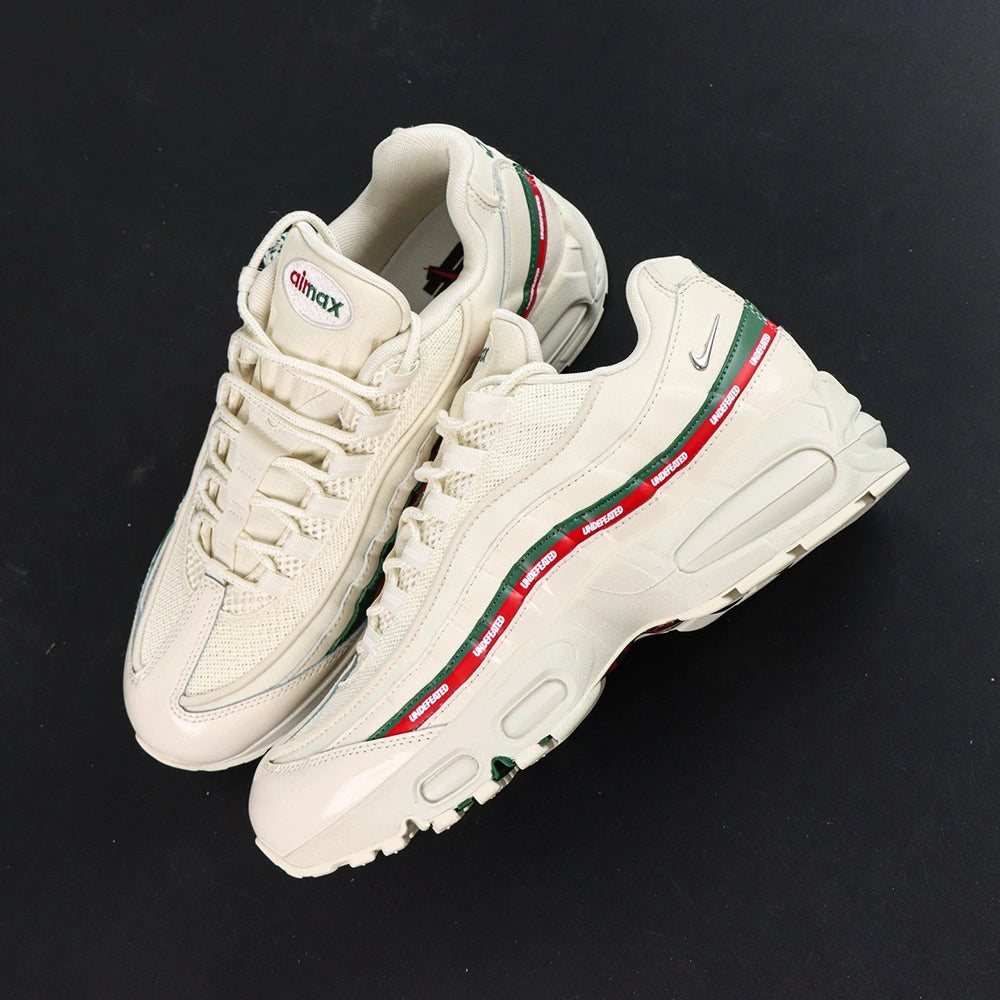 Undefeated x Air Max 95 Sail 2025 - Lateral Profile and Outsole View on Dark Background Showing Sail AM95 Upper Green and Red UNDEFEATED Dual Stripe and Sail White Outsole with Dark Green Square-Block Traction Grid and Red UNDEFEATED Text on Outsole Interior