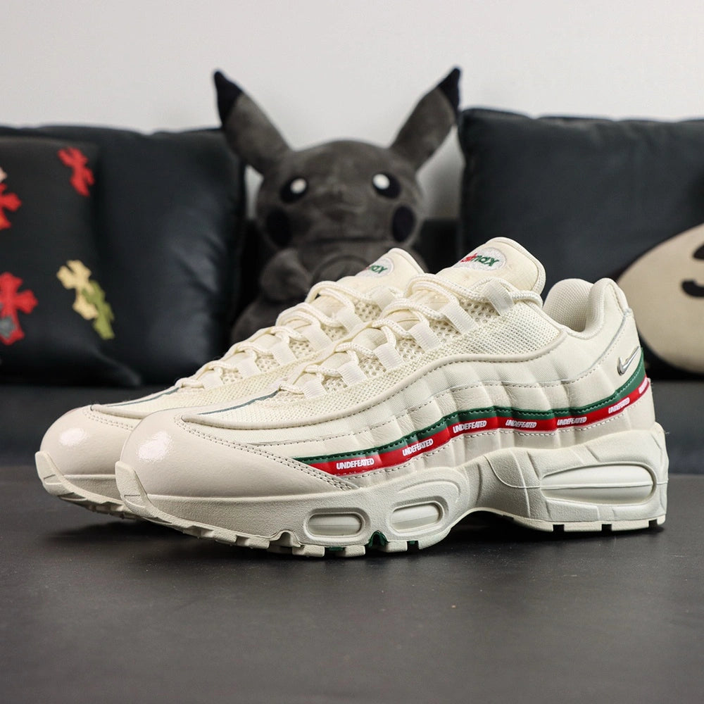 Undefeated x Air Max 95 Sail 2025 - Rear Heel View of Both Shoes on Dark Background Showing Sail AM95 Heel Counter Dark Green Leather Spine Stripe Red UNDEFEATED Ribbon Silver Metal Mini Swoosh on Lateral Heel and NK AIR Text on Tongue
