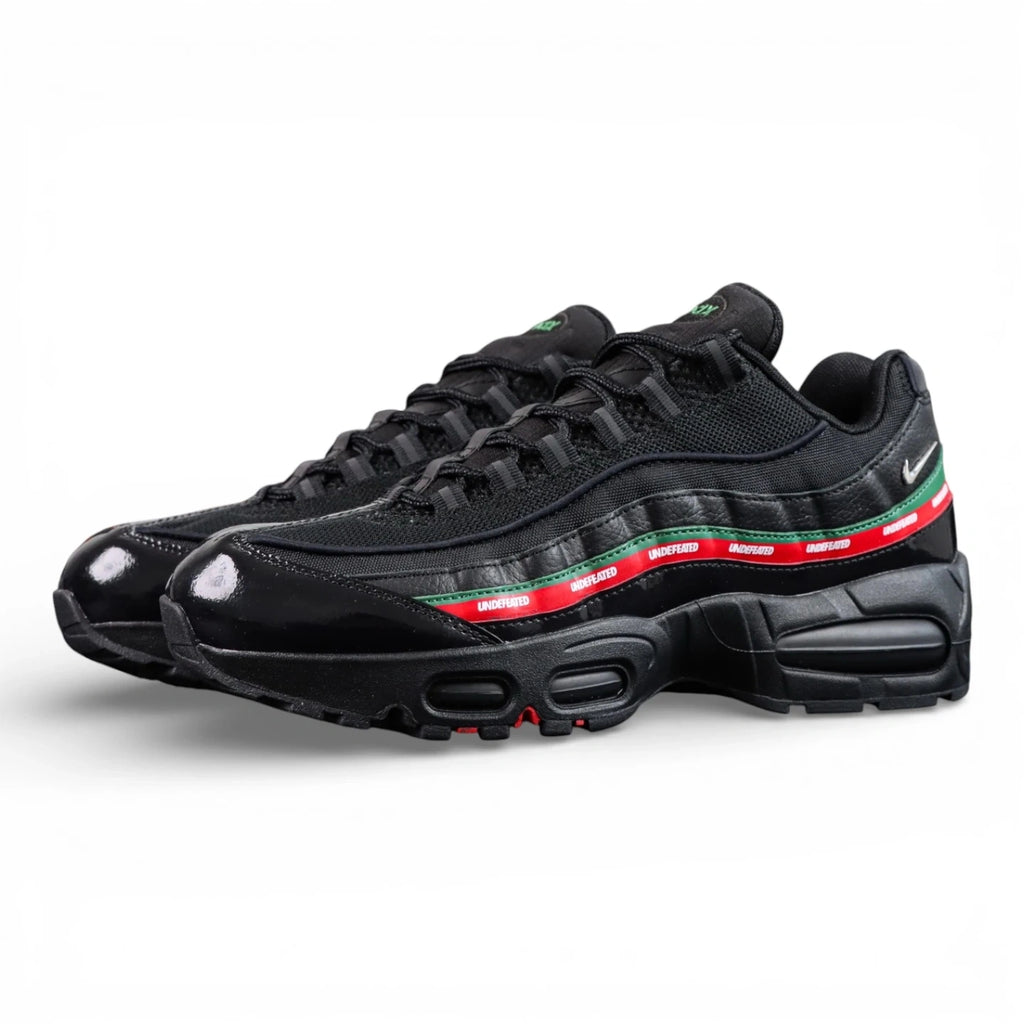 Undefeated x Air Max 95 Black 2025 - Three-Quarter Lateral Top View of Both Shoes on Dark Background Showing Triple Black Patent Leather Mesh and Crinkled Leather AM95 Upper Dark Green Leather Spine Stripe and Red UNDEFEATED Repeat-Text Ribbon