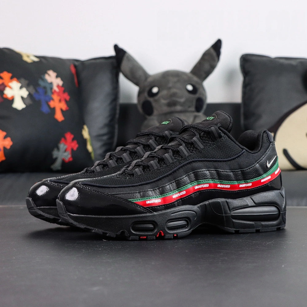 Undefeated x Air Max 95 Black 2025 - Toe Box Close Up on Dark Background Showing Black Patent Leather High-Gloss Mirror Finish Toe Box Black Large-Hole Mesh Quarter Panel and Dark Green Leather Spine Stripe with Red UNDEFEATED Ribbon Below