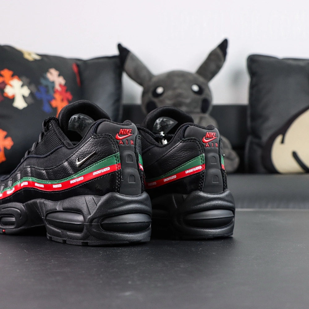 Undefeated x Air Max 95 Black 2025 - Tongue and Ankle Collar Top Down View on Dark Background Showing Black Air Max Oval Badge with Red Air and Green Max Text Black Undefeated Five-Strike Logo on Tongue Interior and Red Textile Ankle Collar Lining