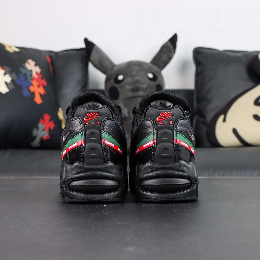Undefeated x Air Max 95 Black 2025 - Lateral Profile and Outsole View with Pikachu Plush on Dark Background Showing Triple Black AM95 Upper Green and Red UNDEFEATED Dual Stripe and Black Outsole with Full Red Square-Block Traction Grid and Green GET THE FUCK OUT Text on Outsole Heel