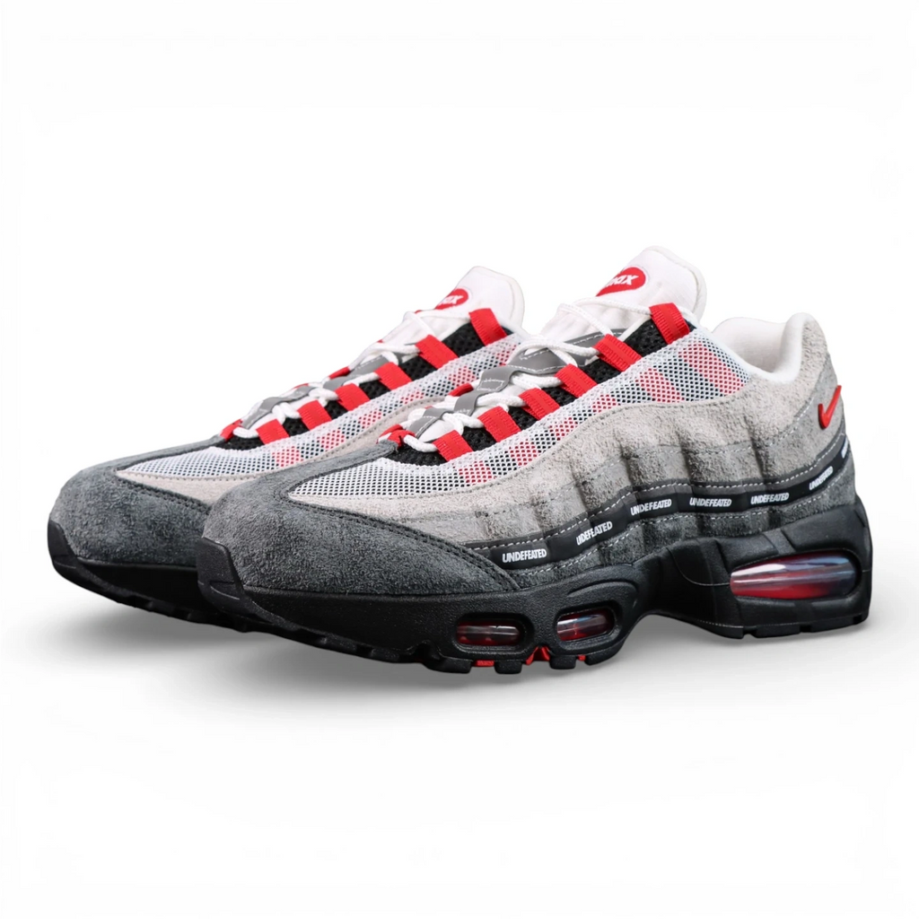 Undefeated x Air Max 95 Bright Crimson 2025 - Three-Quarter Lateral Top View of Both Shoes with White Shoebox on Dark Background Showing White Mesh Grey Suede AM95 Upper Full Black UNDEFEATED Repeat-Text Ribbon Grid on Spine Divider Lines and Red Lace Aglets