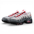Undefeated x Air Max 95 Bright Crimson 2025 - Three-Quarter Lateral Top View of Both Shoes with White Shoebox on Dark Background Showing White Mesh Grey Suede AM95 Upper Full Black UNDEFEATED Repeat-Text Ribbon Grid on Spine Divider Lines and Red Lace Aglets