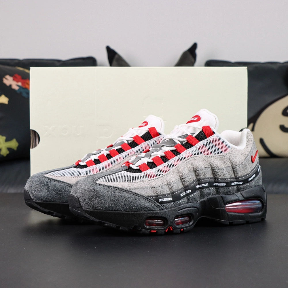 Undefeated x Air Max 95 Bright Crimson 2025 - Rear Heel View of Both Shoes on Dark Background Showing Red NIKE AIR Heel Counter Branding Red Mini Swoosh on Lateral Heel and Crimson AM95 Air Units Visible Through Black Midsole Windows