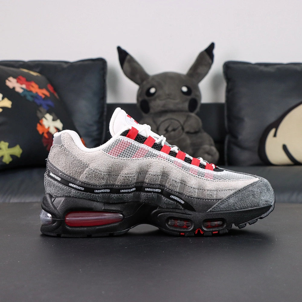 Undefeated x Air Max 95 Bright Crimson 2025 - Toe Box Close Up on Dark Background Showing Dark Grey Suede AM95 Toe Box White Large-Hole Mesh Quarter Panel with Red Mesh Underlayer and Black UNDEFEATED Ribbon Segments on Spine Divider Lines