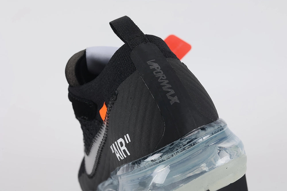 Air VaporMax co-branded Part 2 Black White Orange - Rear View of Both Shoes on White Background Showing Black Woven Heel Pull Tabs with Orange Rectangular Accent White AIR Quotation-Mark Text at Heel Midsole Junction White Co-Branded Text System at Heel Counter Orange Industrial Zip Tie and Frosted Ice VaporMax Air Unit at Heel